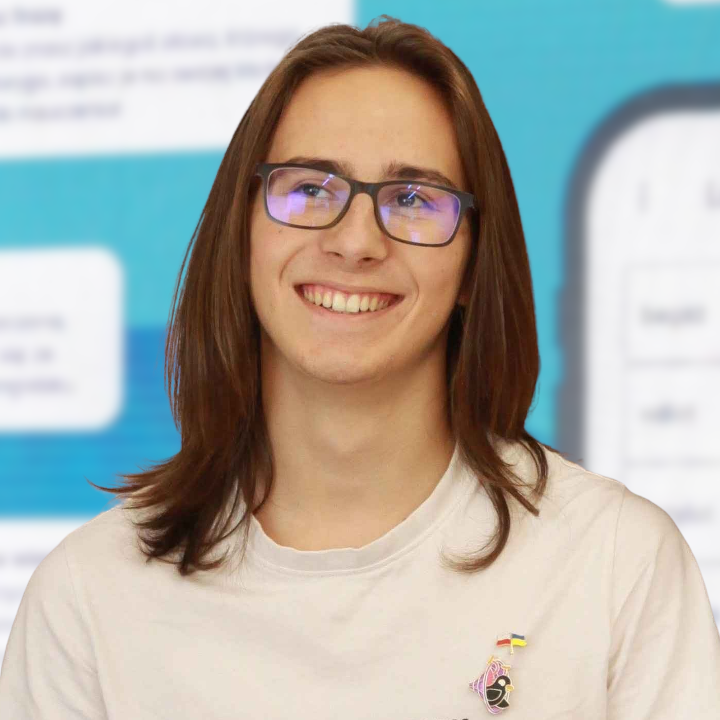 Maciej Saferna smiling, wearing glasses and a white T-shirt with long brown hair. The T-shirt has two metal pins attached: one with artwork from Kurzgesagt featuring two birds in a wormhole and another with the flags of Poland and Ukraine. The blurred background shows the interface of the BorygoAI app.